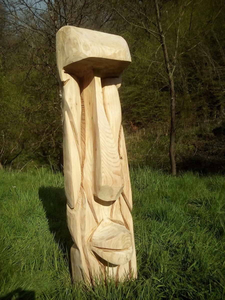 Grande tête sculptée dans un tronc clair, inspirée des statues de l'île de Pâques, debout dans l'herbe.